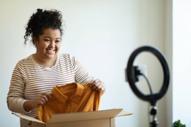 Ünlü blogcu, neşeli, güzel, esmer, kıvırcık, kıvırcık, İspanyol kadın, paket kutusunu kameranın önünde açıyor. Akıllı telefon, takipçilerine yeni bir elbise ya da gömlek gösteriyor.