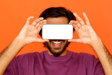 Doğu sakallı, gülümseyen adam gözlerini beyaz ekranlı, yatay akıllı telefonuyla kapatıyor, turuncu stüdyo arka planında bir model. Sosyal medya, cihaz bağımlılığı konsepti