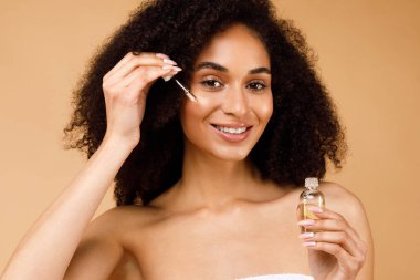 Güzel Afro-Amerikalı kadın, yüz bakımında serum kullanıyor. Güzel bir güzellik ürünü öneriyor. Bej rengi stüdyo arka planında poz veriyor.