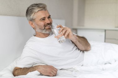 Olumlu, yakışıklı, olgun, gri saçlı, pijama giyen, elinde bir bardak su tutan, sabah uyandıktan sonra su içen ve gülümseyen, ev içi, fotokopi çeken Avrupalı adam.