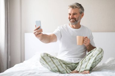 Mutlu, orta yaşlı, beyaz tişört giyen, telefon kullanan, sabah kalktıktan sonra yatakta oturan, akıllı telefon ekranına gülümseyen, kahve içen, telekomünikasyon konsepti olan mutlu, çekici bir adam.