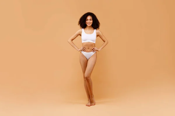 Body contouring and shaping concept. Slender african american lady posing in white underwear over beige studio background, showing her perfect figure and beautiful curves, full length shot