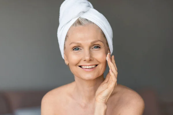 Piękna Seniorka Stosująca Krem Twarzy Uśmiechnięta Kamery Siedząca Sypialni Ręcznikiem — Zdjęcie stockowe