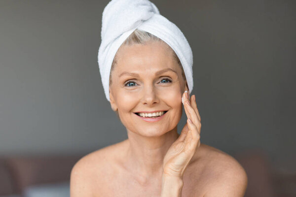 Beautiful senior woman applying face cream and smiling at camera, sitting in bedroom with towel on head after morning shower. Aged lady taking care of her skin, enjoying beauty spa procedure