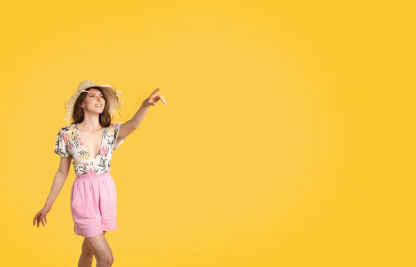 夏季季节性购物 迷人的女士穿着时髦的服装 头戴草帽 在黄色工作室背景上方的复制空间旁边指引方向 大折扣和促销概念 — 图库照片