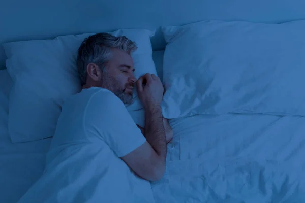 Top view of single handsome grey-haired middle aged man wearing pajamas peacefully sleeping alone in bed at night at home, empty pillow and free space next to him