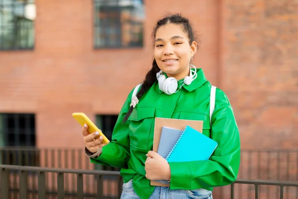 穿着智能手机和练习本的快乐的混血女学生站在大学附近摆姿势 女学生站在外面用手机的画像 — 图库照片
