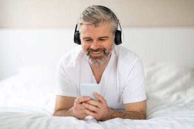 Mutlu, gri saçlı, ev elbiseli orta yaşlı bir adamın portresi. Evde yatıp modern kablosuz kulaklık ve akıllı telefon kullanıyor. İnternetten video içeriğini izliyor.