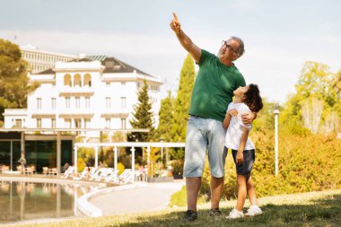 Gülümseyen yaşlı beyaz dede, küçük melez ırk çocuğuna sarıldı, boş alanı gösterdi, boş zamanların tadını çıkardı, dışarıda eğlendi. Dinlenme, hafta sonu ve birlikte parkta tatil, aile ilişkisi.