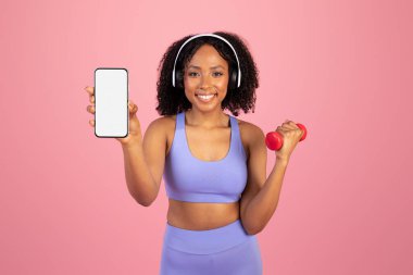 Neşeli milenyum siyahi kadın spor kıyafetleri içinde, kulaklıklı kulaklık pembe arka planda ve stüdyoda boş ekranlı bir telefon gösteriyor. Spor, pilates kondisyonu, vücut bakımı için tavsiye uygulaması