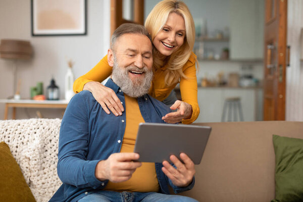 Счастливая пожилая пара наслаждается миром цифровых гаджетов, пользуясь своим компьютером, веб-серфингом и просмотром фильма онлайн в современной гостиной дома. Уход на пенсию и Интернет Веселая концепция