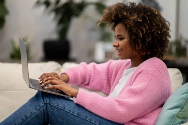 stock image Positive pretty millennial african american lady with bushy hair wearing casual comfy outfit sitting on couch at home, typing on laptop keyboard and smiling, chatting with guys on dating website