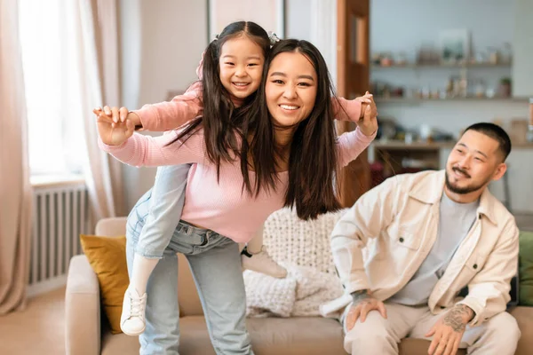 ファミリープレイタイム 赤ちゃんの娘ピギーバックを運ぶ陽気な中国のお母さんは楽しさと遊びを持っています 自宅でソファに座っている間に一緒にポーズ カメラに微笑む 選択的フォーカス — ストック写真