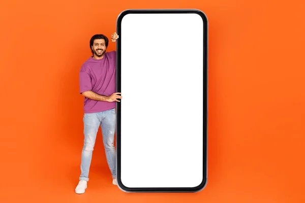 快乐而积极的印度小伙子穿着时髦的休闲装 在网上卖得很好 用白色空白屏幕的大手机摆姿势 模仿橙色工作室的背景 复制空间 — 图库照片