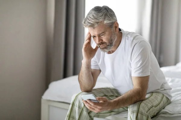 闷闷不乐的中年男人穿着睡衣坐在床上 用手机碰头 读坏消息 收到奇怪的信息或电子邮件 复制空间 — 图库照片