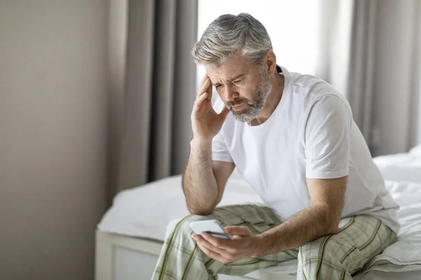 stock image Upset unhappy handsome middle aged man wearing pajamas sitting on bed at home, using cell phone and touching head, reading bad news, got weird message or email, copy space