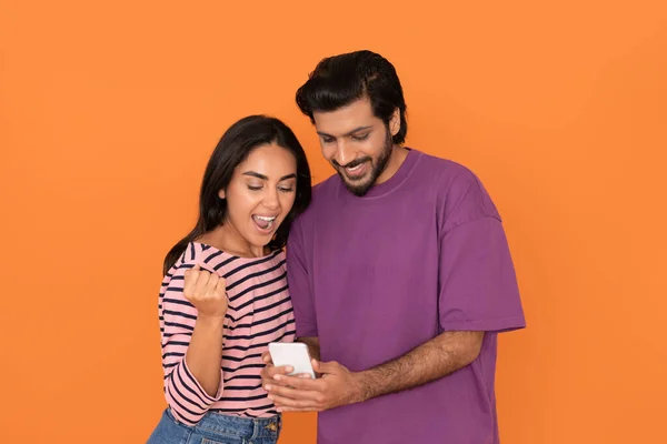 感情的な幸せな若い東部の恋人男性と女性でカジュアル衣装で携帯電話を使用して ガジェット画面を見て オレンジのスタジオの背景にジェスチャー 株式取引 コピースペース — ストック写真