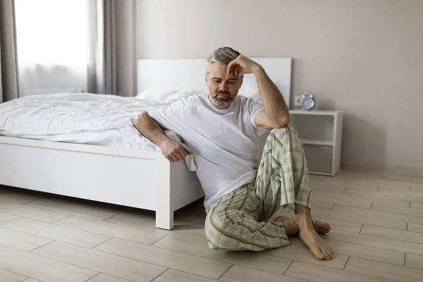 stock image Upset pensive middle aged grey-haired handsome man wearing pajamas sitting on floor by bed at home, touching head, suffering from hangover or anxiety, copy space. Permacrisis, midlife crisis