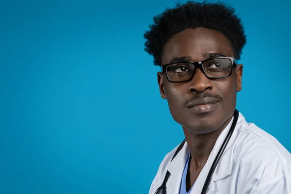 健康専門家 魅力的な若いですアフリカ系アメリカ人男で医学コートの医師身に着けています眼鏡脇に見て クローズアップスタジオショット上の青の背景 テキストや広告のためのコピースペース — ストック写真