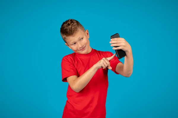 快乐的未成年男孩站在蓝色工作室的背景下使用手机 快乐的未成年男孩在智能手机上浏览互联网或自拍 享受现代技术 复制空间 — 图库照片
