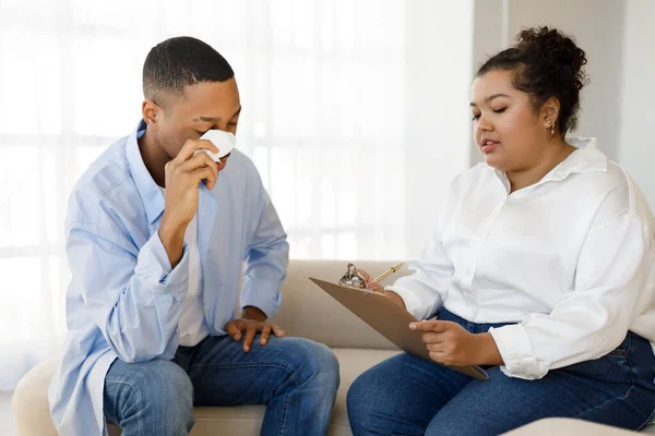 Mental health concept. Distressed upset young black man patient sitting on couch, holding napkin, crying during session with psychotherapist hispanic lady plus size, clinic interior, copy space
