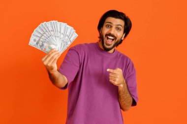 Heyecanlı, zengin, zengin, yakışıklı, mor tişörtlü, para basan banknotlar ve buruşukluklar gösteren, turuncu stüdyo geçmişinde izole edilmiş, ticaret yapan, online kumar oynayan, zengin ve yakışıklı bir adam.