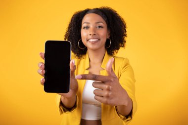 Yenilik davet etmek. Elinde boş ekranla cep telefonunu işaret eden mutlu zenci kadın güzel bir online teklif sunuyor, maket yapıyor, sarı stüdyo arka planında poz veriyor.