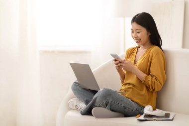 Günümüz modern teknolojileri gündelik hayatta. Mutlu, genç, Asyalı, günlük kıyafetler giyen, evdeki koltukta oturan, dizüstü bilgisayar kullanan, akıllı telefon, tablet, kulaklık takan güzel bir kadın.