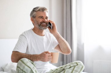 Neşeli, gülümseyen, orta yaşlı, beyaz saçlı, sakallı bir adam evde oturmuş, kahve içiyor, telefonla konuşuyor, reklam için fotokopi odasına bakıyor, pijama giyiyor.