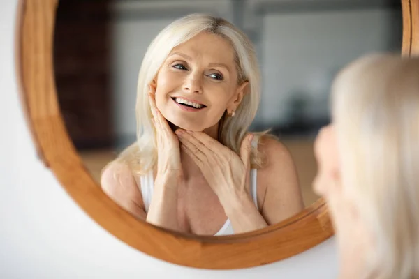 快乐迷人的老妇人看着镜子 摸着松软的脖子 享受着家里的反射和抗衰老的日常护肤 — 图库照片