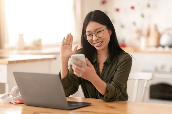 スマートフォンの画面とジェスチャーを見て ノートパソコンの前にキッチンデスクに座って眼鏡をかけている陽気な若い可愛いアジアの女性は ビジネスパートナーとビデオ通話を持っている スペースをコピー — ストック写真