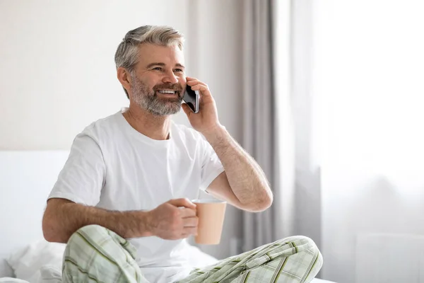 快乐地微笑着 英俊的中年白发苍苍的男人坐在床上 喝咖啡 打电话 看广告的复制空间 穿睡衣 — 图库照片