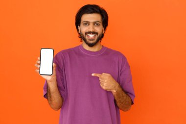 Bin yıllık gündelik kıyafetli çekici Hindu adam güzel bir online teklif sunuyor, cep telefonunu beyaz ekran ile tutuyor, maketi gösteriyor, turuncu stüdyo arka planında izole edilmiş.