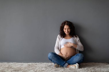 Bebek beklentisi. Gri duvarın yanında otururken hamile karnını kucaklayan güzel genç bir kadın, mutlu bir anne, karnına sevgiyle bakıyor, hamileliğin tadını çıkarıyor, boşluğu kopyalıyor.