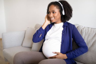 Bin yıllık gülümseyen Afro-Amerikan kadın kulaklıklı koca göbekli müzik dinliyor, oda içinde rahatlıyor. Ses uygulaması, boş zaman, doğum, çocuk beklentisi, annelik.