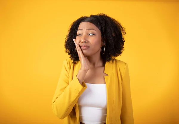 Sad young african american lady in casual suffering from toothache, feeling pain and touching cheek isolated on yellow background, studio shot. Health problems, dentistry, caries and sensitive teeth