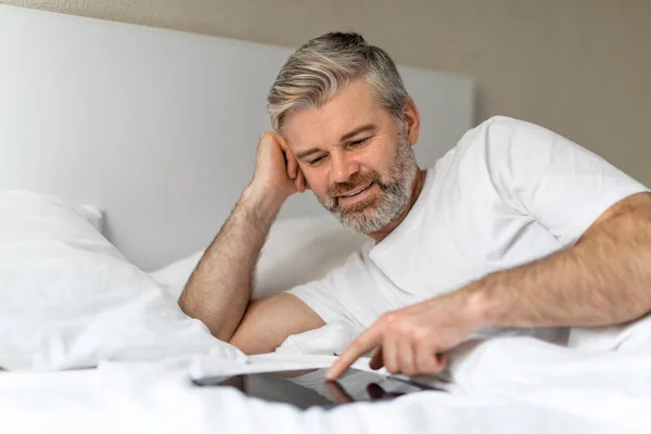 中年男人穿着白色的T恤躺在床上 用现代Pc数码记事本 触摸桌子空白屏幕 在网上滚动 复制空间 — 图库照片