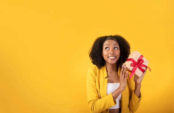 Ideas for gift. Excited glad black woman holding gift box and looking at blank space isolated on yellow background, studio shot. Woman thinking of anniversary, Christmas and New Year present