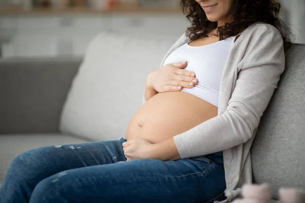 幸せな笑顔妊娠中の女性がリビングルームでソファに座って 若い期待の母親が腹を抱いて 彼女の赤ちゃんのバンプを見て 今後の母親を楽しんで クローズアップ撮影 — ストック写真