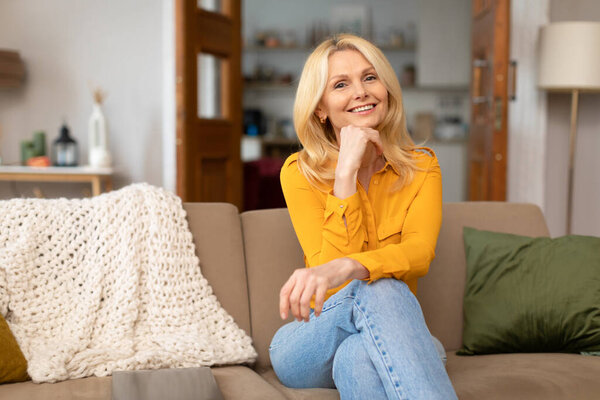 Нестареющая красота. Великолепная пожилая блондинка позирует дома на диване, улыбаясь в камеру трогательным лицом. Портрет уверенной в себе зрелой леди, выражающей позитивные эмоции в гостиной