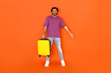 Neşeli, mutlu, şık, şık Hintli adam turuncu stüdyo arka planında sarı bavul taşıyor. Turist, güzel bir seyahat anlaşması için fotokopi alanının yanında duruyor.