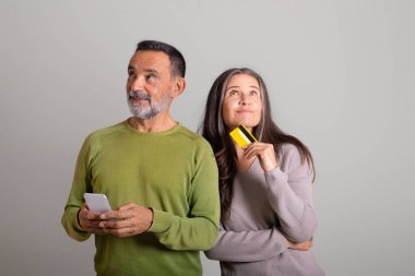 Akıllı telefonlu dalgın, beyaz, yaşlı bir adam ve gri stüdyo arka planında bir düzen kuran kredi kartı olan bir kadın. İlişki, online alışveriş, satış, indirim ve para iadesi.