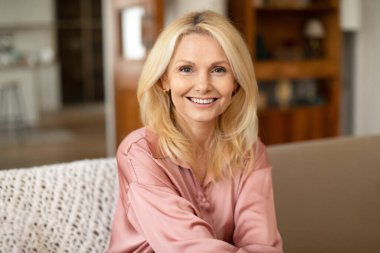 Portrait Of Blonde European Mature Woman Posing Smiling To Camera Sitting On Sofa At Home. Lady Resting On Cozy Couch In Modern Living Room Indoor. Ageless Beauty And Elegance Concept clipart