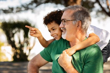 Olumlu küçük melez çocuk yaşlı beyaz dedeye sarılıyor, fotokopi alanını işaret ediyor, dışarıda eğleniyor. Hafta sonu dinlenme ve parkta tatil, aile ilişkisi.
