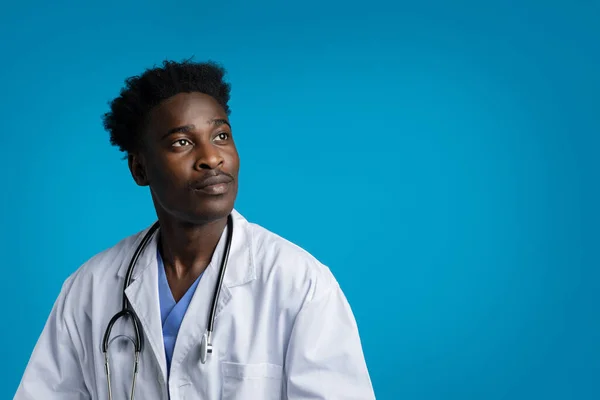 有兴趣的年轻黑人穿着医疗外套 脖子上戴着听诊器 医生看着蓝色工作室背景的复制空间 医护人员的专业教育 — 图库照片