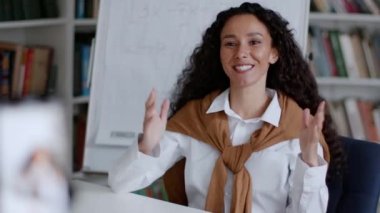 Kişisel eğitim blogu. Genç pozitif orta yaşlı bayan öğretmen akıllı telefondan video kaydı dersi alıyor, abonelere el sallıyor ve gülümsüyor, çekim takip ediyor, yavaş çekim