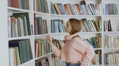 Geleneksel bilgi kaynağı. Kütüphanedeki kitaplıklarda kitap arayan genç pozitif kadın öğrenci, üniversitedeki sınavlara hazırlanıyor, çekim takibi, yavaş çekim, boş alan