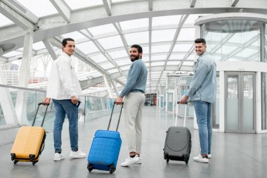 Uçuş zamanı. Seyahat bavullu üç mutlu yolcu modern havalimanı kalkış terminalinde çağrı posizyonunu bekliyor, kameraya gülümsüyor. Tam Uzunluk