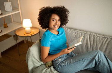Ders çalış, planlama günü evde. Neşeli genç siyah kıvırcık kadın kitap okuyor, kanepeye oturuyor, boş zamanların ve eğitimin tadını çıkarıyor, hobisi var ve akşamları oturma odasında dinleniyor.
