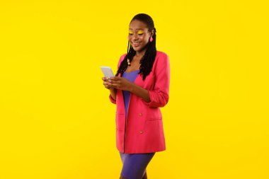 Yeni Uygulama İlanı. Gülümseyen Siyah Kadın Akıllı Telefon Kullanıyor, Cep Telefonu Uygulamaları Çevrimiçi İletişimin keyfini çıkarıyor Sarı Stüdyo Arkaplanında Ayakta Duran Sohbetler. Dijital Bağlantı ve Eğlence